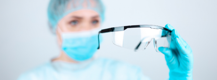 Female in face mask, gown and gloves holding a pari of safety glasses out in front of her.